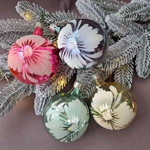 May include: Four vintage glass Christmas ornaments in pink, silver, teal, and gold, each with a frosted daisy design. The ornaments are arranged on a frosted evergreen branch, creating a festive holiday scene.