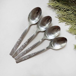 Puede incluir: Un juego de cuatro cucharas de metal plateado dispuestas en fila diagonal. Cada cuchara presenta un cuenco ovalado pulido y un mango plano decorado con un patrón floral en relieve que consiste en hojas y rosas en la sección inferior.
