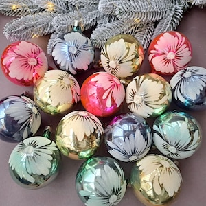 May include: A collection of colorful Christmas ornaments. The ornaments are round, with a floral design in white. The colors include pink, blue, green, and gold. The ornaments are arranged on a surface with frosted greenery in the background.