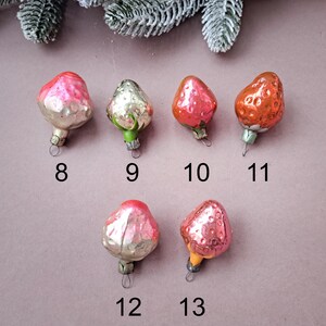 May include: A collection of vintage strawberry-shaped Christmas ornaments. The ornaments are in various shades of red, pink, and silver, with green or yellow stems. Each ornament has a small metal loop for hanging, and numbers 8, 9, 10, 11, 12, and 13 are printed below them.