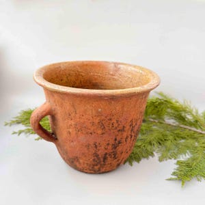 Op de afbeelding: Een rustieke, terracotta-kleurige keramische beker met een handvat. De beker heeft een brede, afgeronde body en een uitlopende rand. Donkere vlekken geven textuur aan de buitenkant. Een takje groen loof ligt naast de beker, tegen een witte achtergrond.
