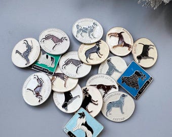 May include: A selection of vintage dog breed pins in various shapes. The pins feature detailed illustrations of different dog breeds, with text in a foreign language. The pins are primarily silver and white, with accents of black, blue, and brown.