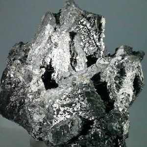 Könnte beinhalten: Ein Cluster aus metallisch grauen, silberfarbenen und schwarzen Kristallen. Die Kristalle sind in einer unregelmäßigen, gezackten Form angeordnet.