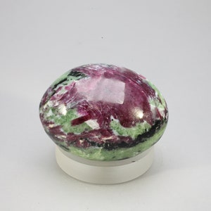 Op de afbeelding: Een gladde, ovale groene en roze edelsteen met zwarte en witte insluitsels. De edelsteen is gepolijst en heeft een glanzende afwerking.