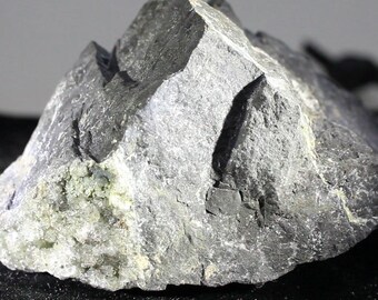 May include: A large, dark gray, rough-textured rock with a few small, light green crystals embedded in it.