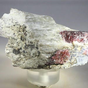 May include: A raw mineral specimen featuring a large, translucent white crystalline matrix with embedded deep red, faceted-looking crystals. The stone has a rough, uneven texture with visible grey and black mineral inclusions throughout the surface.