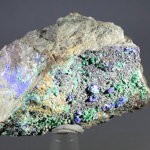May include: A close-up of a mineral specimen featuring a mix of colors and textures. The rock has a rough, gray exterior with a band of dark gray, speckled with vibrant green and patches of blue. Yellow and brown hues are also visible.