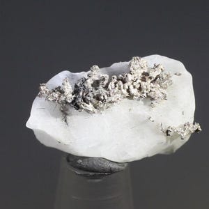 May include: A close-up of a natural mineral specimen. The piece features a white, rounded base with a cluster of silver-colored, crystalline structures on top. The mineral sits on a dark gray base against a gray background.