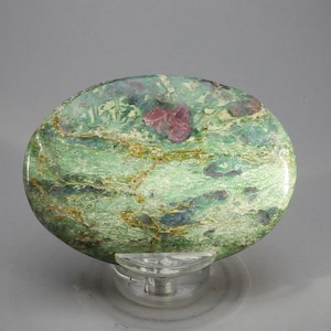 May include: A polished, oval gemstone, predominantly green with gold streaks and patches of red and blue. The stone is displayed on a clear stand. The gemstone's unique colour variations are highlighted by the polished surface, creating a striking visual effect.