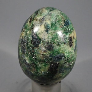 May include: A polished, egg-shaped stone with a mottled pattern of green, black, and white. The stone has a smooth, glossy surface and is set against a neutral background. The overall appearance suggests a decorative or collectible item.
