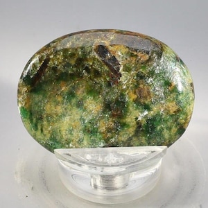 May include: A polished, oval-shaped gemstone with a mix of green, yellow, and brown hues. The stone is set on a clear, rounded display stand, showcasing its natural patterns and textures. The gemstone's surface is smooth and reflective.