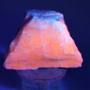 May include: A raw mineral specimen exhibiting intense fluorescent orange coloration under ultraviolet light. The stone has a rough, crystalline texture with a jagged, uneven surface and a blocky, somewhat pyramidal shape, resting on a clear stand.