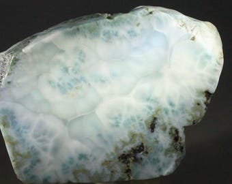 May include: A polished piece of larimar gemstone, showcasing a blend of white, light blue, and teal hues. The stone's surface displays a unique pattern of swirling colors and textures, with dark spots adding contrast. The overall shape is irregular.