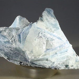 May include: A crystalline mineral specimen with a light blue and white color palette. The mineral has a rough, jagged texture and appears to be composed of layered formations. The specimen is displayed on a metallic base, showcasing its natural beauty.