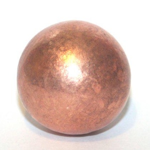 Puede incluir: Una esfera de color cobre con una superficie texturizada. La bola metálica tiene un tono marrón rojizo cálido con reflejos más claros, lo que sugiere un acabado pulido. La forma esférica es prominente sobre un fondo blanco.