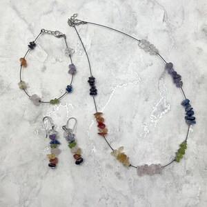 May include: A set of rainbow gemstone jewelry featuring a necklace, bracelet, and earrings. The necklace and bracelet are made with black string and feature small, colorful gemstones. The earrings are dangle earrings with a cluster of gemstones.