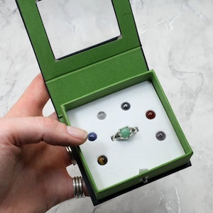 May include: A silver ring with a green gemstone center, displayed in a green and black box with other small gemstones. The box is open, revealing the ring and gemstones. The ring has a floral design around the gemstone.