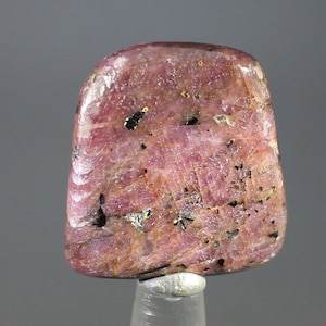 May include: A polished, rectangular gemstone with a rounded top. The stone is a mix of pink and reddish-brown hues, with black and gold flecks throughout. It is mounted on a clear, cylindrical stand.