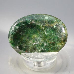 May include: A polished, oval-shaped gemstone with a vibrant green hue, resting on a clear, curved display stand. The stone's surface displays a mottled pattern of lighter and darker shades of green, creating a natural, organic appearance.