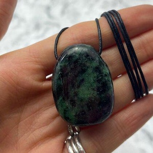 May include: A polished, oval-shaped zoisite stone pendant featuring deep forest green and dark black mottled patterns. The smooth, rounded gemstone is suspended from a thin, black cord necklace that loops through the top of the stone setting.