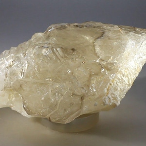 Puede incluir: Un espécimen mineral translúcido de forma irregular con un tono amarillo pálido. La superficie tiene una estructura cristalina en capas con fracturas visibles y una textura ligeramente rugosa. El mineral se asienta sobre una base blanca, con una luz suave y difusa.