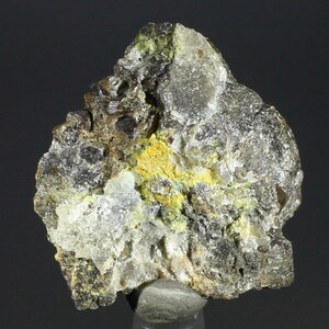 Puede incluir: Primer plano de una roca gris oscura con depósitos minerales amarillos y verdes. La roca tiene una superficie rugosa y desigual.