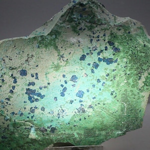 May include: A close-up of a rough-textured rock formation with a mix of green, blue, and off-white colors. The surface is speckled with dark blue clusters, and the overall appearance suggests a natural mineral specimen.