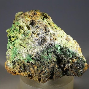 Puede incluir: Un espécimen mineral en bruto, de forma irregular, con una mezcla de colores. La roca presenta áreas de verde brillante, amarillo, marrón y negro, con algunas manchas cristalinas blancas. La superficie tiene un aspecto texturizado y poroso.