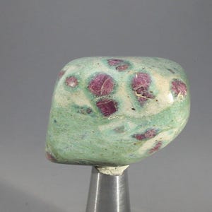 May include: A polished gemstone with a mottled green and white surface, displaying striking red inclusions. The stone is mounted on a silver-coloured metal stand, highlighting its natural beauty and unique colour variations.