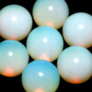 May include: Seven smooth, round, white opal cabochons with a slight blue hue. The opals are arranged in a circle on a black background.