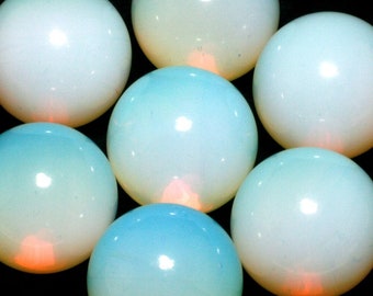 May include: Seven smooth, round, white opal cabochons with a slight blue hue. The opals are arranged in a circle on a black background.