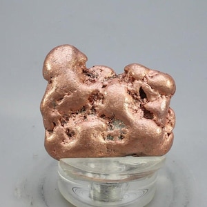 May include: A raw, metallic copper nugget with a distinct reddish-brown hue and an irregular, textured surface. The specimen features rounded protrusions and deep, porous crevices throughout its structure, showcasing a natural, unrefined appearance.