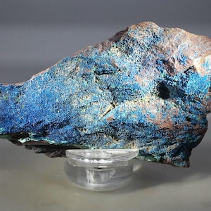 May include: A raw mineral specimen featuring a vibrant, crystalline blue surface texture contrasted against a rough, earthy brown rock base. The mineral is displayed resting on a clear, circular acrylic stand, showcasing its natural, jagged form.