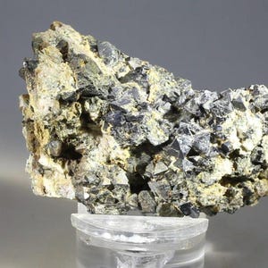 May include: A raw mineral specimen featuring a cluster of dark, metallic, multifaceted crystals embedded within a rough, light-colored, textured matrix. The crystalline structure displays sharp, geometric edges and a reflective, charcoal-gray surface.