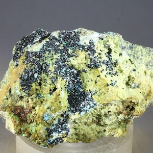 Puede incluir: Primer plano de un espécimen mineral con una superficie rugosa y texturizada. La roca muestra una mezcla de colores, incluyendo amarillo-verdoso, azul-negro oscuro y toques de marrón. El mineral parece ser cristalino en algunas áreas.