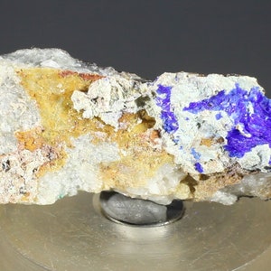 May include: A close-up of a rough, gray rock with patches of blue, silver, and yellow minerals. The rock is sitting on a silver stand.