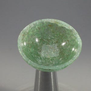 May include: A small, round, light green glass bowl with a textured surface. The bowl is supported by a silver-colored metal stand. The bowl's interior is visible, and the glass appears to have small inclusions.
