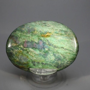 May include: An oval-shaped polished gemstone featuring a vibrant green base colour with marbled textures and distinct inclusions of deep pink and purple mineral deposits throughout the surface, resting on a clear, curved plastic display stand.