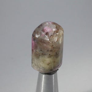 May include: A polished gemstone with a rounded top and a cylindrical shape. The stone features a mix of colours, including pink, brown, and cream, with a translucent quality. It is mounted on a silver metal stand.