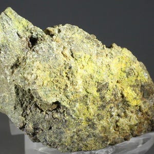 Puede incluir: Primer plano de una formación rocosa rugosa y texturizada. La roca presenta una mezcla de colores, incluyendo amarillo, verde y marrón. La superficie es irregular con pequeñas grietas y variaciones de color, sugiriendo un espécimen mineralógico o geológico.