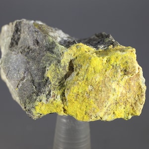 Puede incluir: Una roca amarilla y gris con una textura rugosa. La roca tiene un depósito mineral amarillo brillante en un lado.