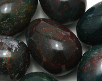 May include: Close-up of several polished bloodstone eggs. The eggs have a dark green base color with red and brown inclusions. The smooth, glossy surface reflects light, highlighting the natural patterns and variations in the stone.