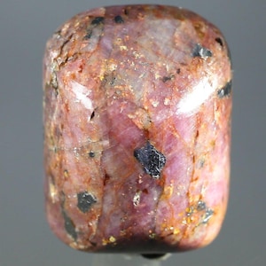 Peut inclure: Une pierre précieuse cylindrique polie, avec un mélange de teintes roses, rouges et brunes. La pierre présente des inclusions sombres et irrégulières et une forme légèrement arrondie. La surface a un aspect naturel et texturé.