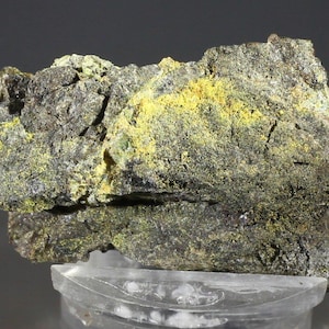 Puede incluir: Primer plano de una formación rocosa rugosa y texturizada. La roca es principalmente de color gris oscuro con parches de amarillo brillante y verde. La roca se asienta sobre una superficie clara y transparente, mostrando su aspecto natural y en bruto.