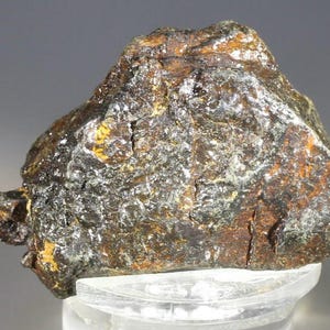 Puede incluir: Un espécimen mineral en bruto de forma irregular, con un aspecto oscuro y moteado. La superficie muestra una mezcla de marrón oscuro, negro y toques de oro y plata, con un acabado ligeramente texturizado. El mineral se encuentra sobre una base transparente.