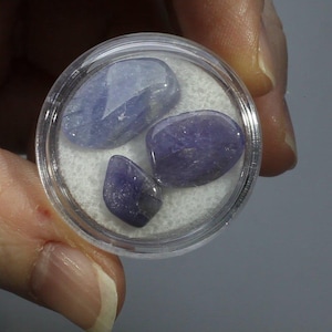 May include: A small, clear plastic container holding three polished, oval-shaped, blue-purple gemstones. The gemstones are displayed on a white, textured surface. The container is held by a hand.