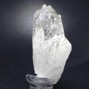 Puede incluir: Un grupo de cristales de cuarzo transparente con una parte superior puntiaguda y una superficie rugosa y texturizada. El cristal está montado sobre un soporte transparente sobre un fondo oscuro. Las facetas del cristal reflejan la luz.
