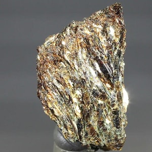 May include: A raw mineral specimen featuring a jagged, irregular shape with a highly reflective, metallic surface. The stone displays a complex texture of deep brown, bronze, and golden hues, with sharp, fibrous striations running vertically.
