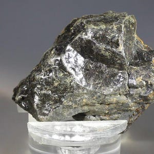 Może przedstawiać: Ciemny, nieregularny kształt okazu mineralnego o szorstkiej, teksturowanej powierzchni. Skała ma głęboki czarny kolor z odcieniami zieleni i brązu. Znajduje się na przezroczystym, okrągłym stojaku.