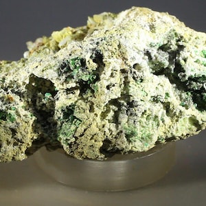 Puede incluir: Primer plano de un espécimen mineral con una superficie rugosa y texturizada. La roca presenta una mezcla de colores, incluyendo verde claro, verde oscuro y amarillo, con áreas negras y blancas. El mineral se encuentra sobre una base clara y redondeada.